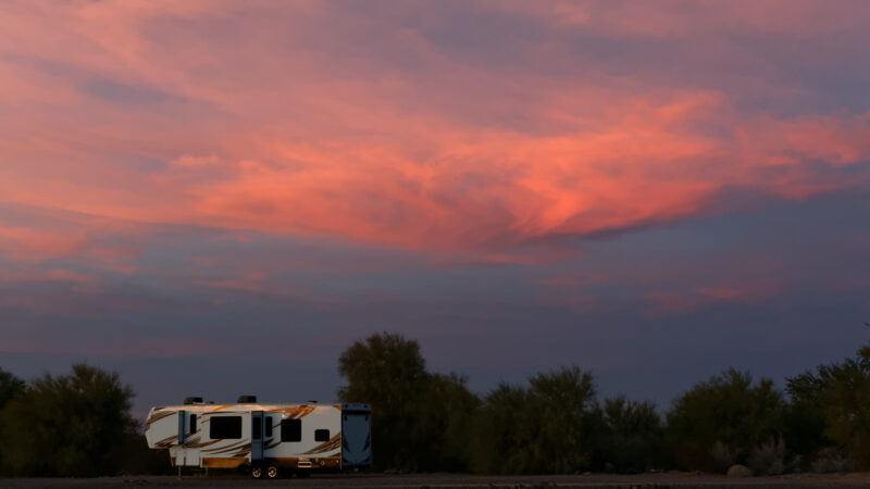 A fifth wheel RV is longterm boondocking in a desert with a pink sunset in the sky.