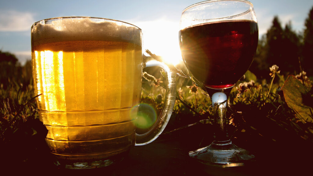 A large glass of beer and glass of wine shine in the sun against grass and flowers.