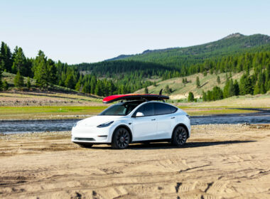 a Tesla Model Y parked in the sand with water and mountains behind it
