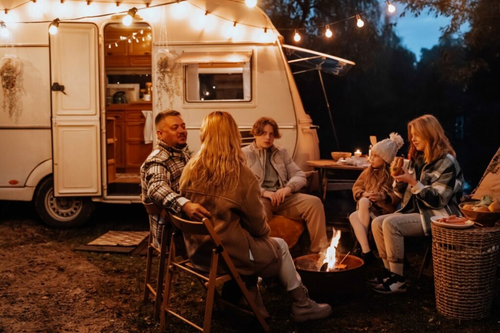 Family sits around the fire outside of their travel trailer with string lights illuminating the campsite.