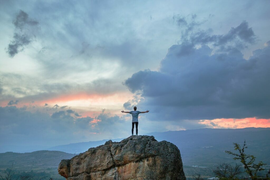 A beautiful sky of blue and orange set behind a man standing atop a rock formation stretching his arms out.