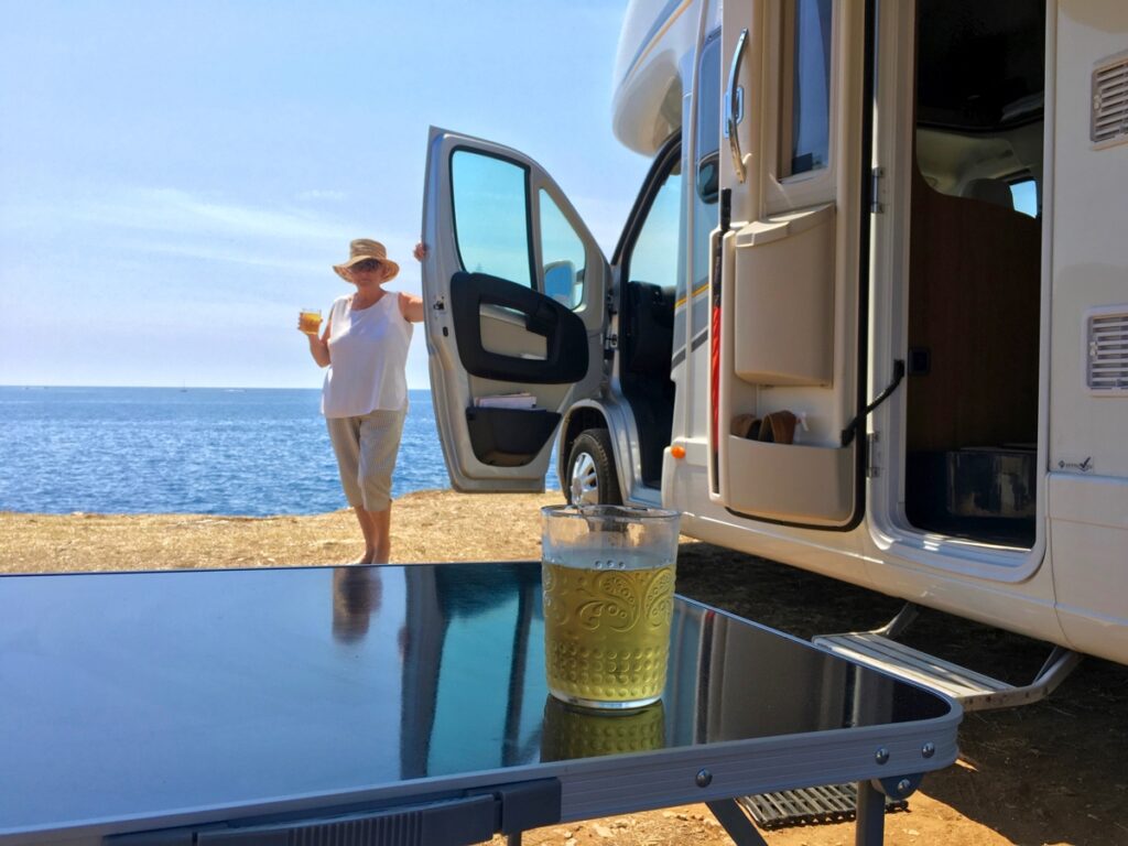 Woman holds a drink and stands next to her motorhome parked on the beach with the ocean in the background.