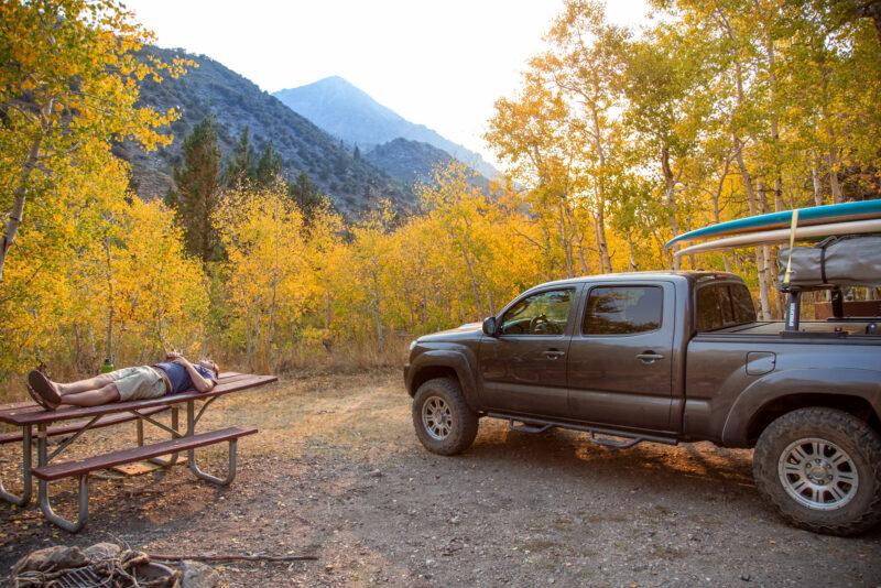 A camper naps on a picnic table while camping in the autumn forest in California with his truck and surfboard.