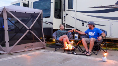 A couple cheers outside there RV while sitting in camping chairs and surrounded by fun RV accessories.