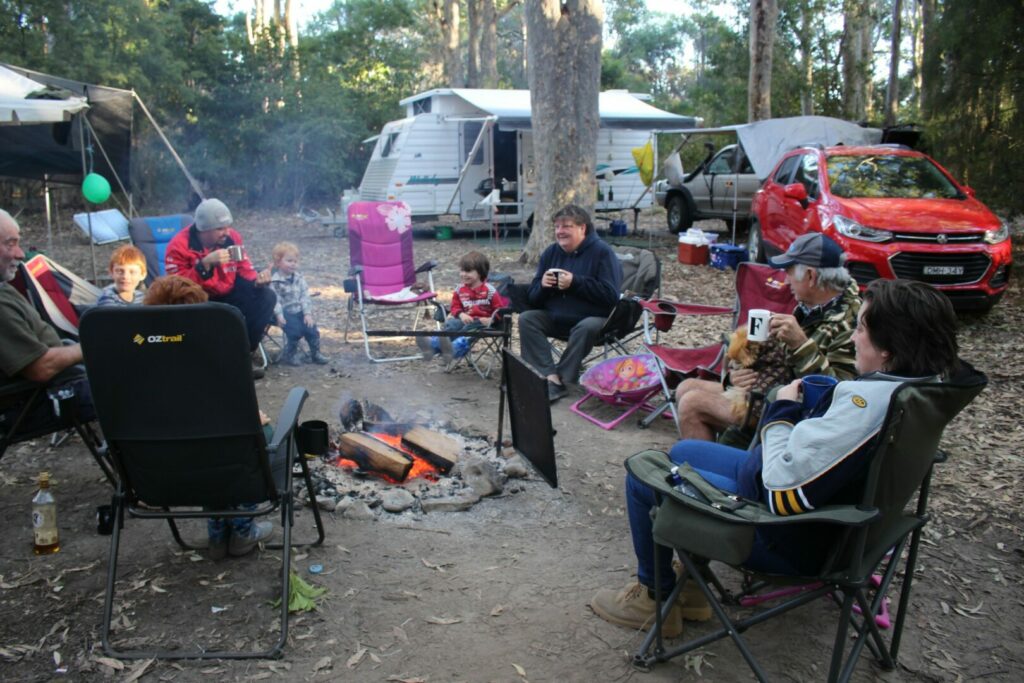 Families sit around a campfire at their campground drinking hot beverages out of mugs.