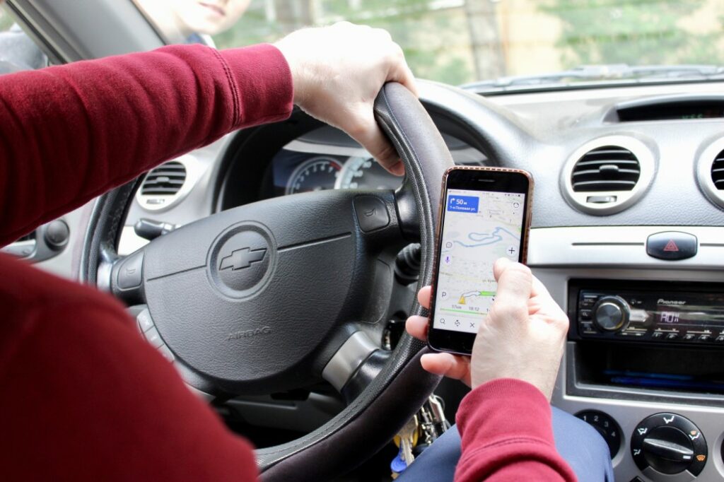 Close up of a man holding his phone with a GPS app open in one hand and grasping the steering wheel of his vehicle with the other.