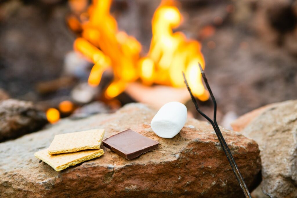 Graham crackers, chocolate, a marshmallow and a roasting stick laying on a stone while a fire roars out of focus in the background.