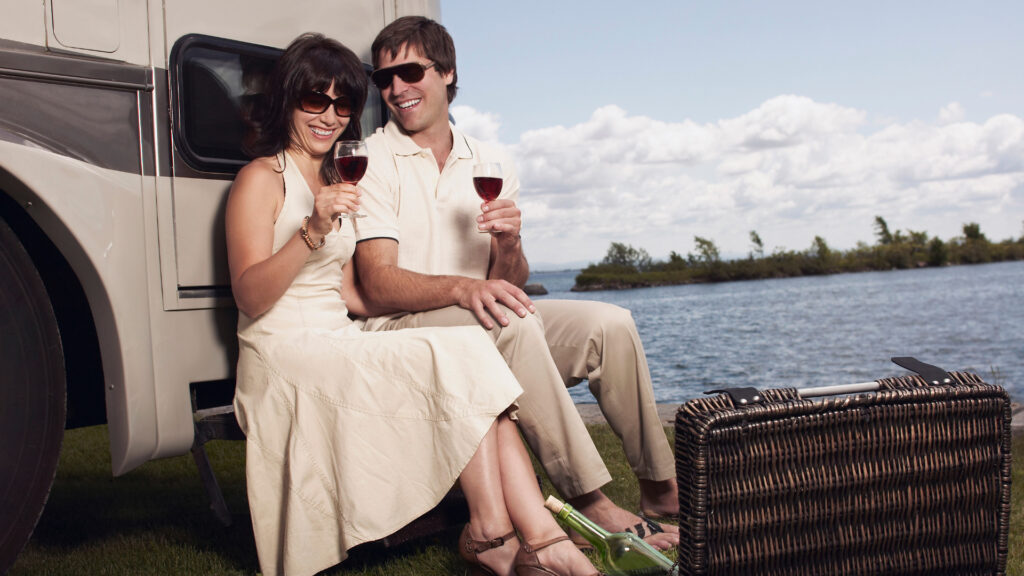 A couple smile happily and enjoy a glass of wine along some water outside of their RV.