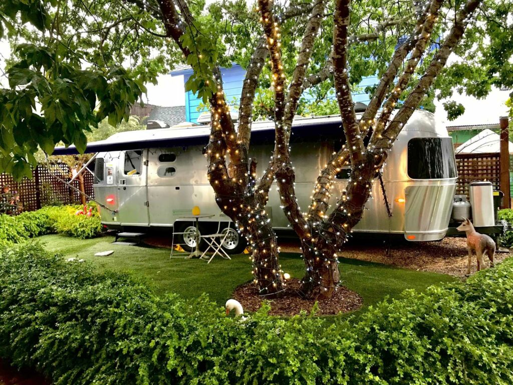 A large airstream travel trail parked in a fancy campsite with string lights wrapped around the trees and manicured landscaping.