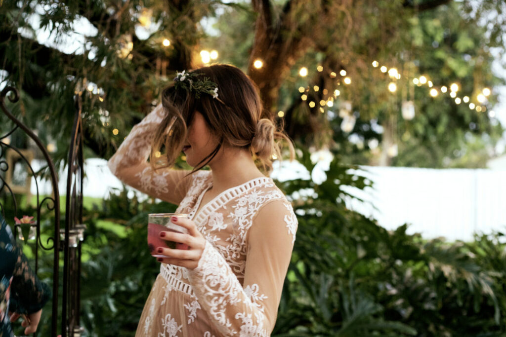 A woman in a lacey white dress dances carefree outside with a drink in her hand.