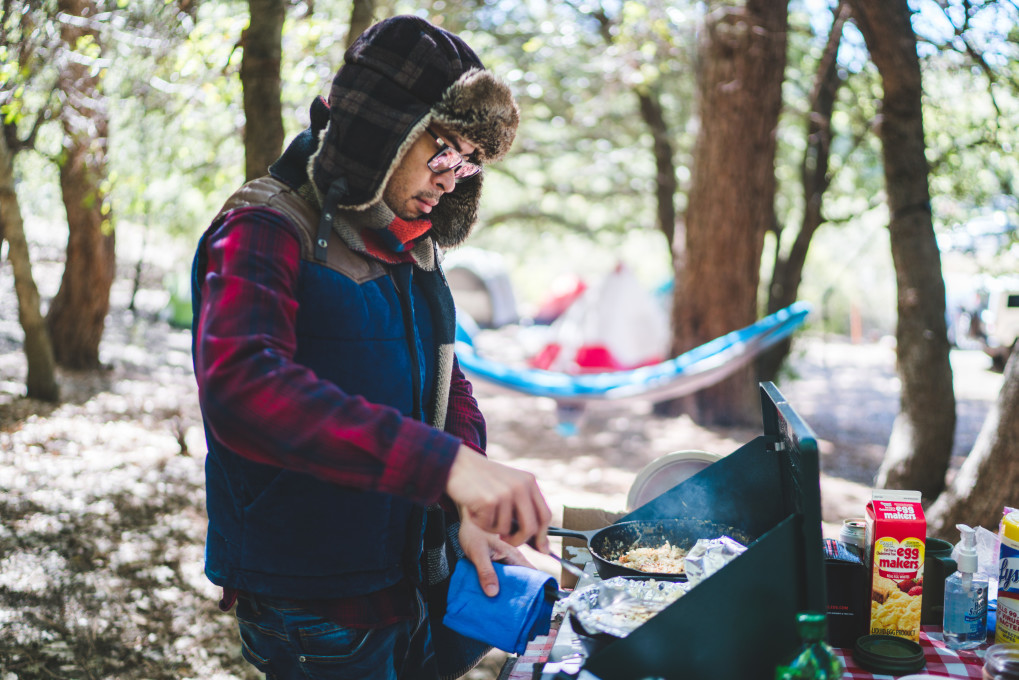 Man cooking an outdoor camp breakfast on a griddle wearing a warm winter hat, vest, and flanner.
