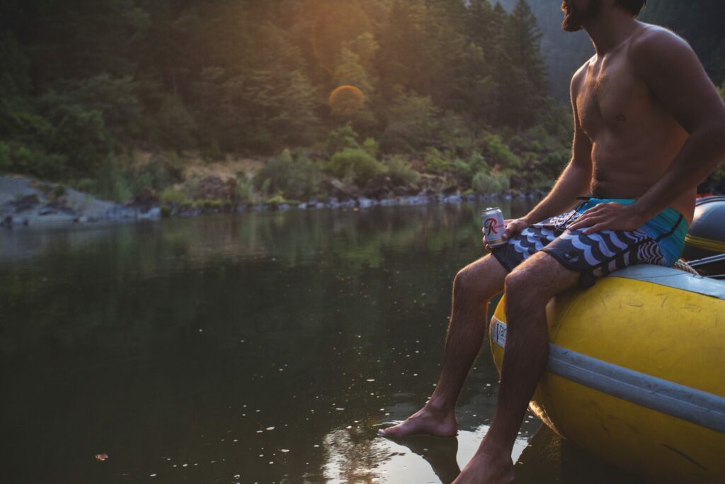A man in a bathing suit sitting on the edge of a blow up boat on a river holding a cheap beer