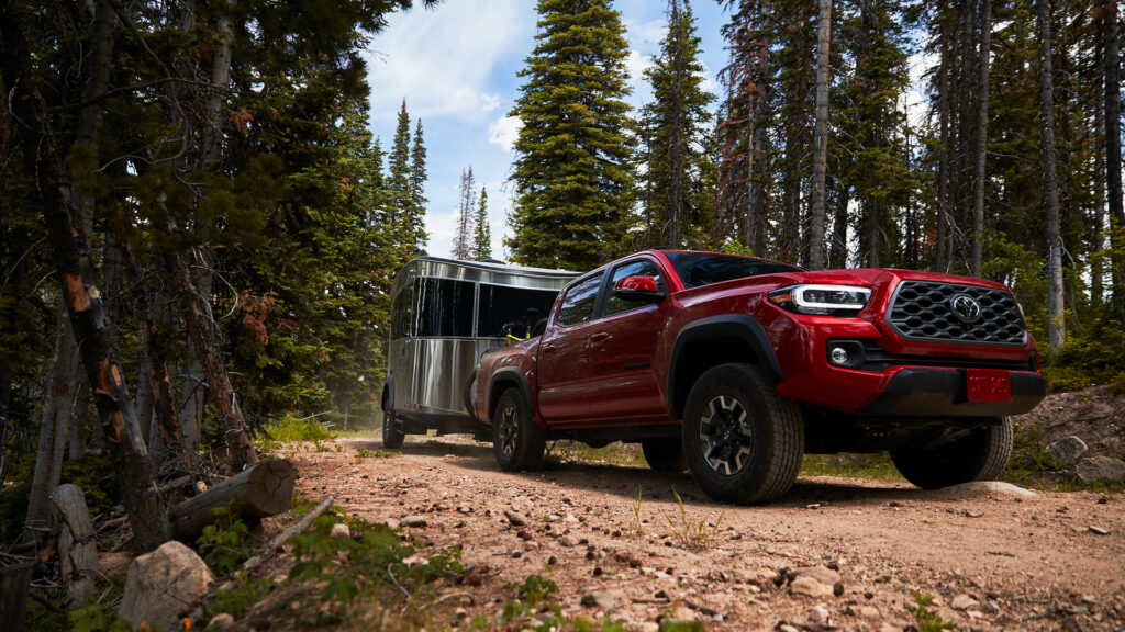 A red truck tows the airstream basecamp trailer down a narrow dirt road in the forest.