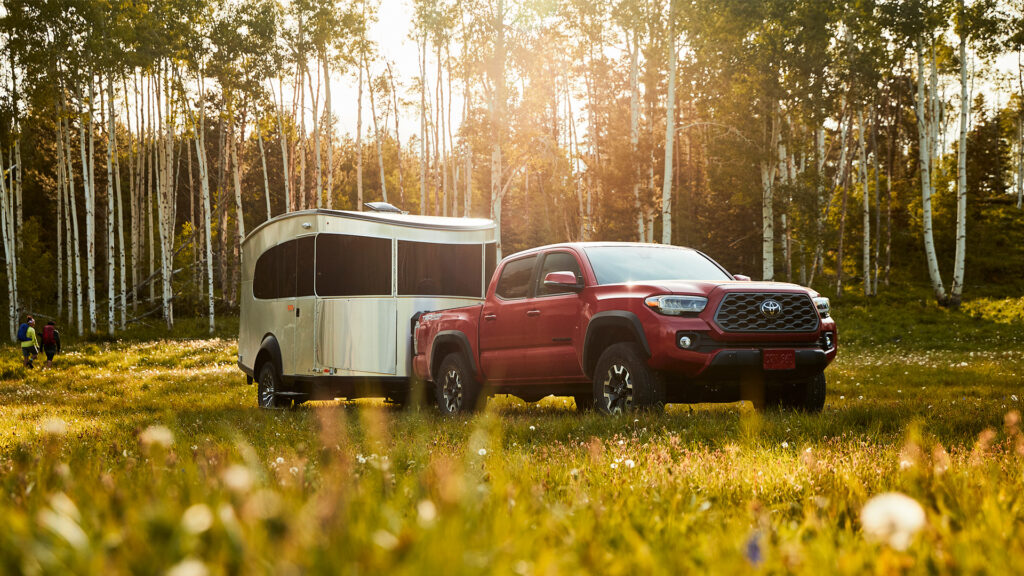 A red truck tows the Airstream Basecamp travel trailer through a grassy meadow surrounded by trees.