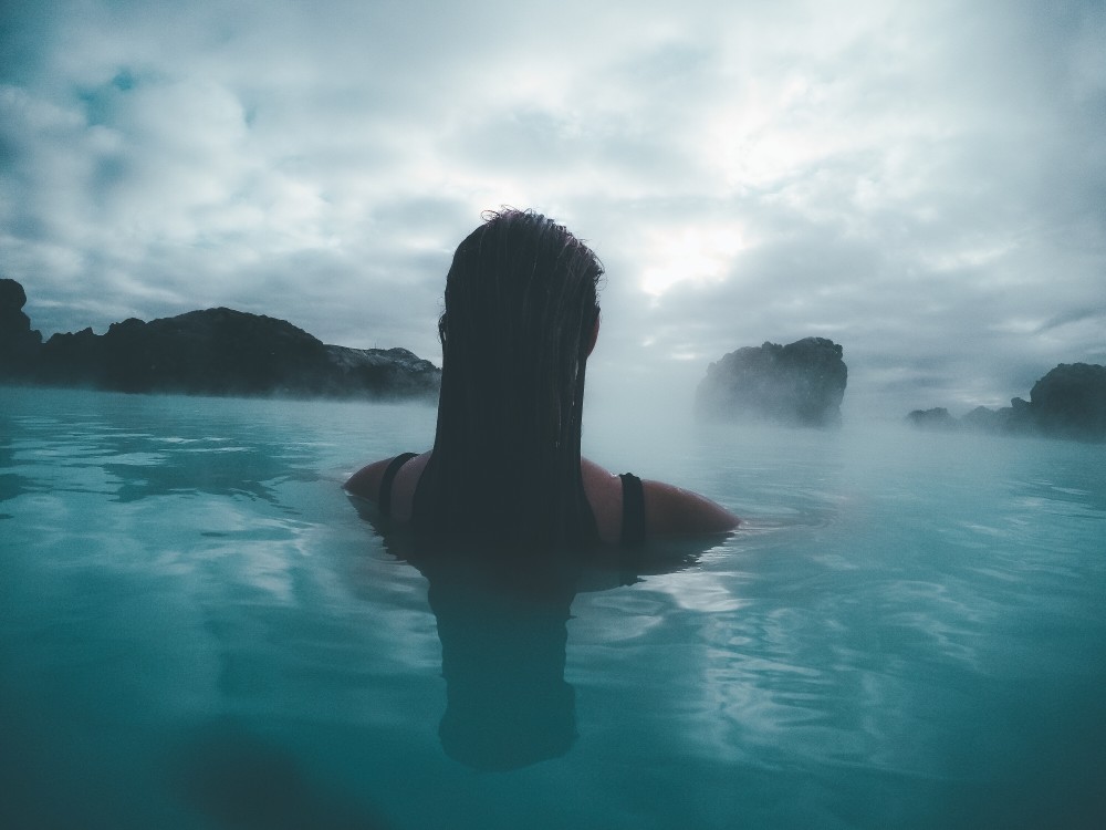 Silhoutte of the back of a woman's head and shoulder while she's soaking in a large hot spring on a cool cloudy day