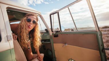 Young woman with curly blonde hair and a head band playfully sticks her tongue out at the camera as she hangs out of a vintage van door.