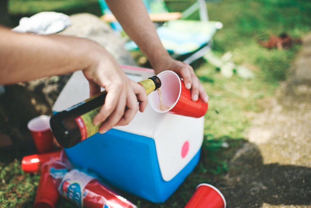 A cold beer is poured into a red plastic cup with a cooler and more cups strewn on the ground in the background of the campsite.