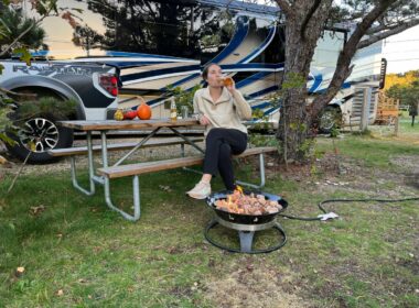 A young woman drinks a beer on a bench near a firepit and her RV and truck are in the background.
