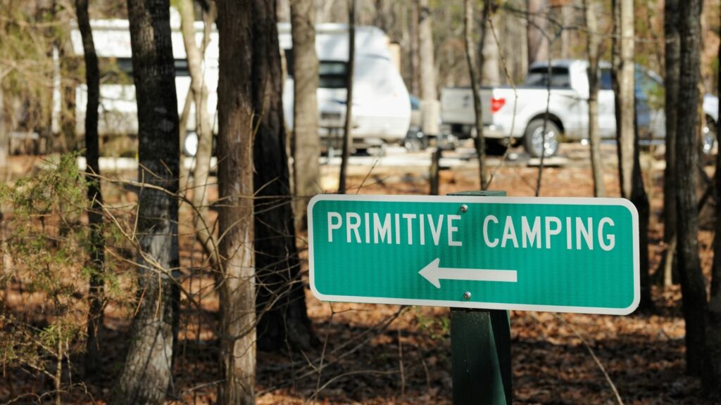 Green sign pointing to an area for primitive camping with a truck and travel trailer beyond the trees in the distance.