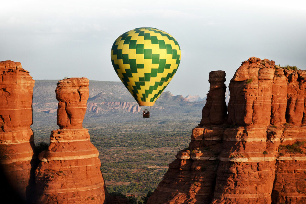 A green a yellow hot air balloon floats through tall red rock formations in Sedona, Arizona.