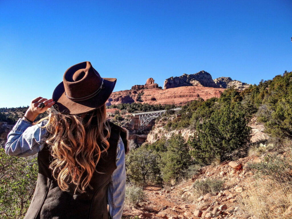 From behind, a woman wearing a vest, flannel, and hat hiking through the landscape of Sedona, Arizona.
