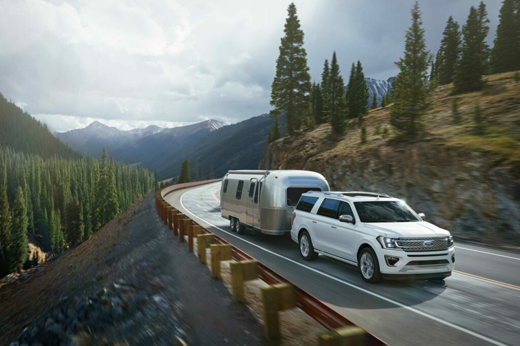 White Ford Expedition tows an Airstream travel trailer up a mountainside covered in pines.