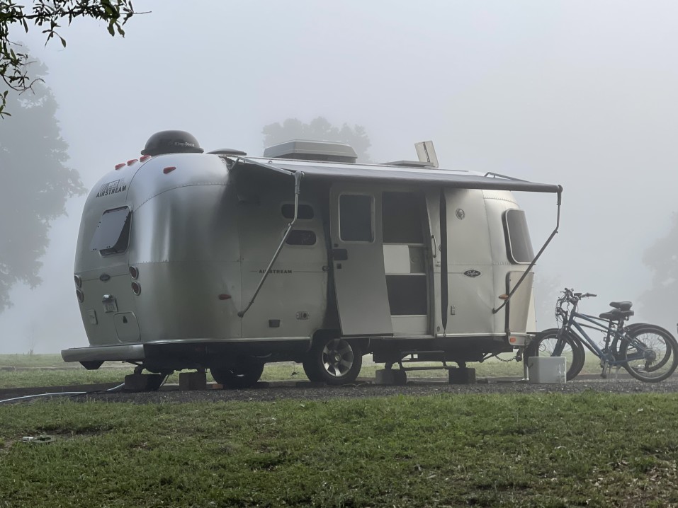 An Airstream travel trailer with the awning stretched out at a foggy morning campsite.