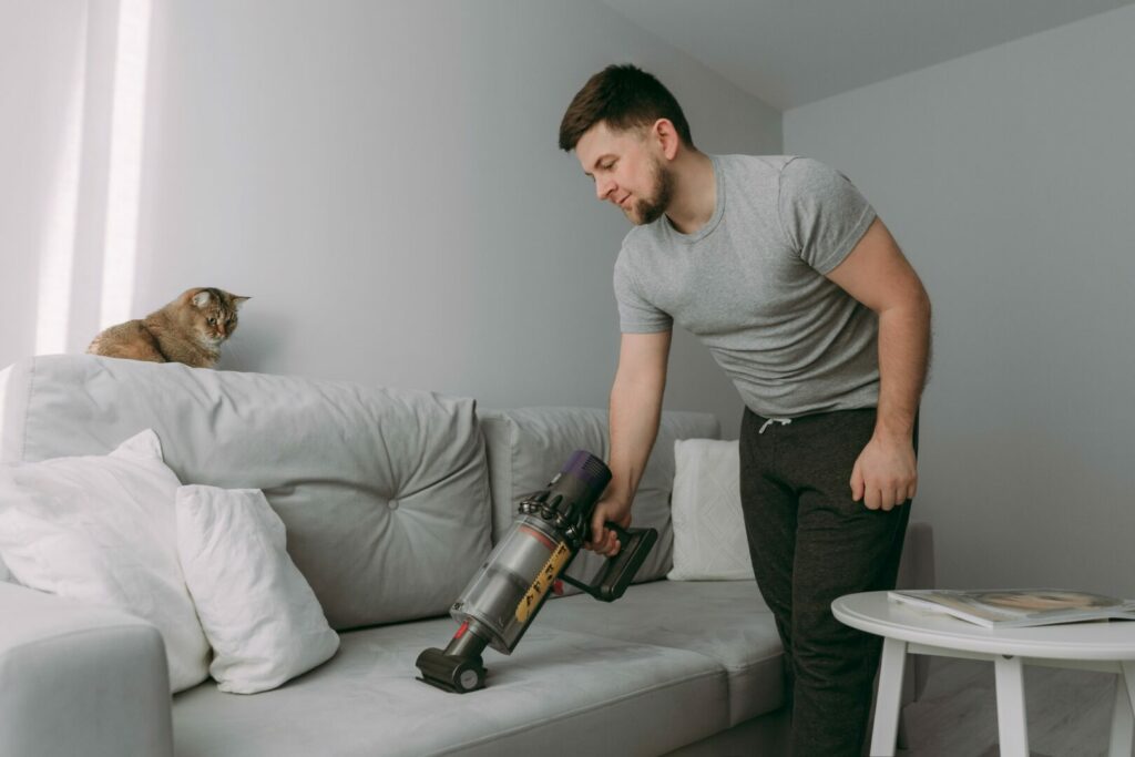 Man using a hand vacuum to clean the couch while a chonky orange cat watches with interest.