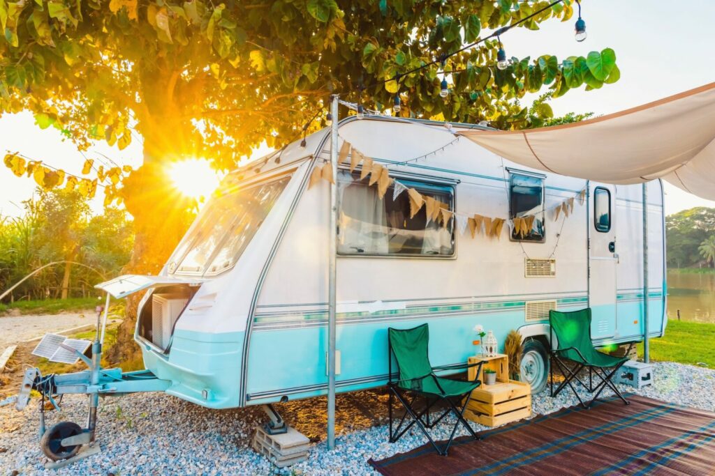 Camping chairs placed outside cozy retro travel trailer Caravan under tree before sunset near the river in peaceful countryside.