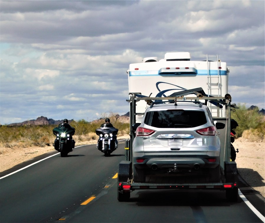 Motorhome tows a silver SUV down a two lane road as motorcycles pass.