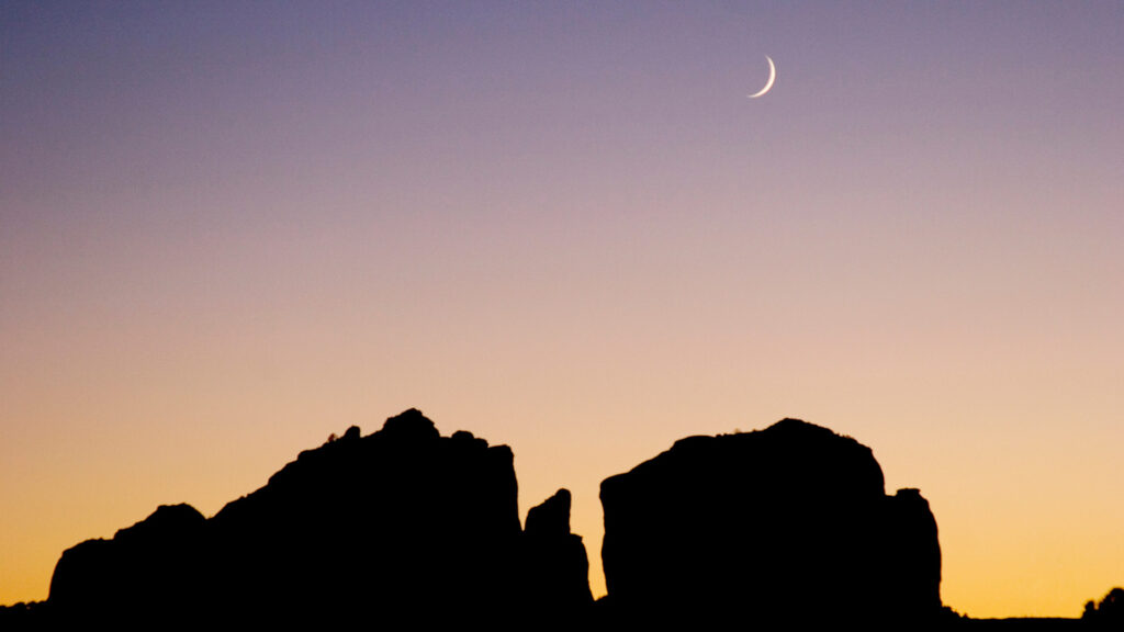 A sunset sky over rocks in Sedona, AZ with a crescent moon shining overhead.
