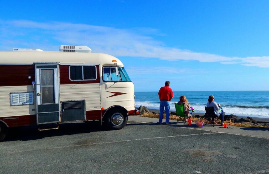 A family sets up their camping chairs on the pavement by the ocean with their motorhome parked next to them.