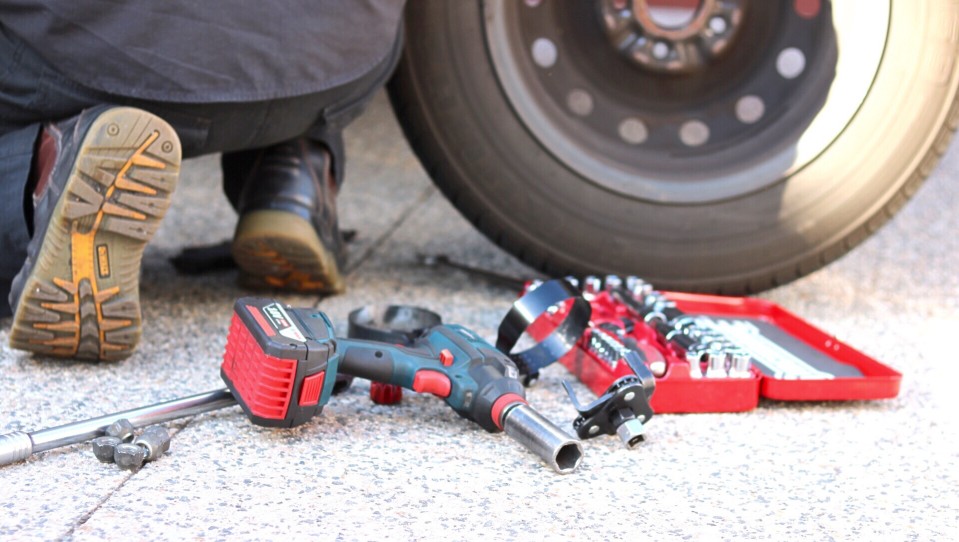 Tools and a tool box lay on the ground behind a person kneeling down by the tire of a vehicle.