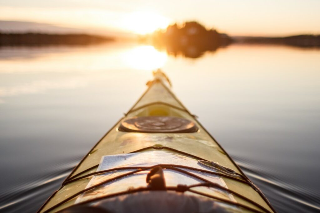 The front of a kayak points towards the sunrise on a calm lake.