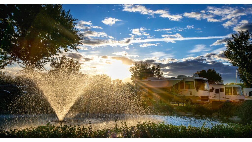 Sun stream through a water fountain at an RV resort with motorhomes parked in the grass.