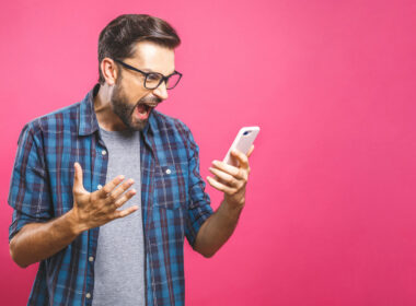 A man with glasses and a bear holds his phone up and yells at it angrily.