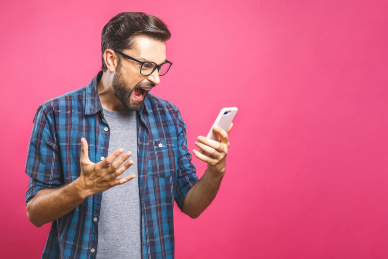 A man with glasses and a bear holds his phone up and yells at it angrily.