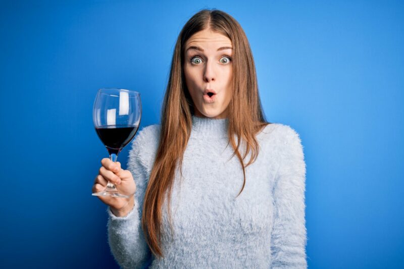 Young beautiful redhead woman drinking glass of red wine over isolated blue background scared in shock with a surprise face, afraid and excited with fear expression