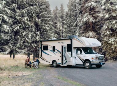 Two people sit outside their class C RV in early winter to enjoy the snow covered trees.