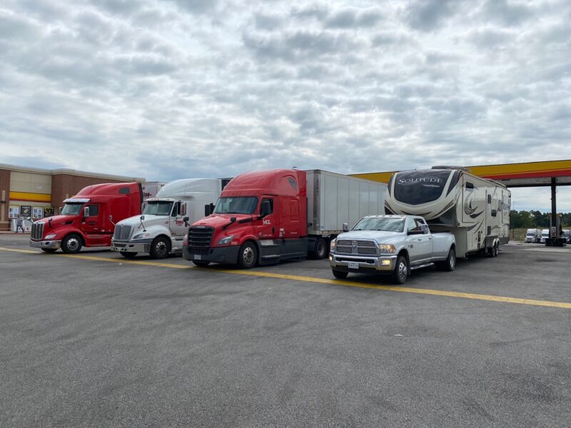 A fifth wheel rv is being towed by a large ram truck and parked next to big rig trucks at a truck stop.