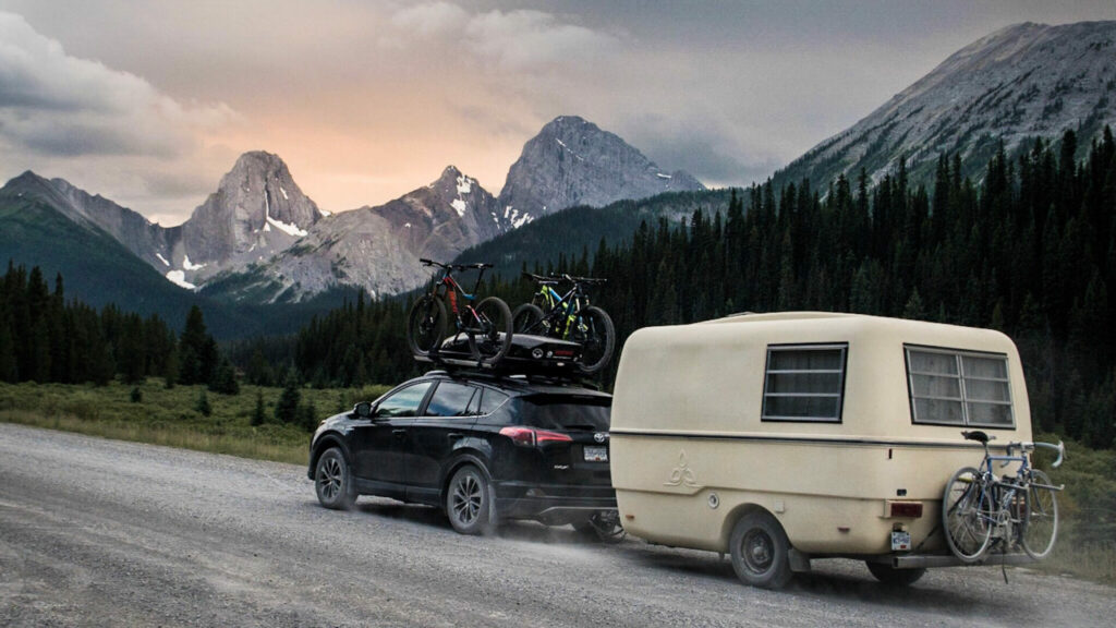 A small crossover SUV pulls a travel trailer down a gravel road with an amazing view of rocky mountains in the distant.