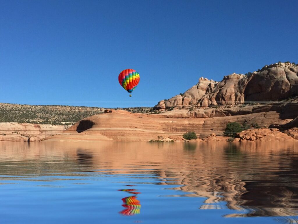 A colorful rainbow hot hair balloon low in the sky, reflecting in a lake in Moab Utah.