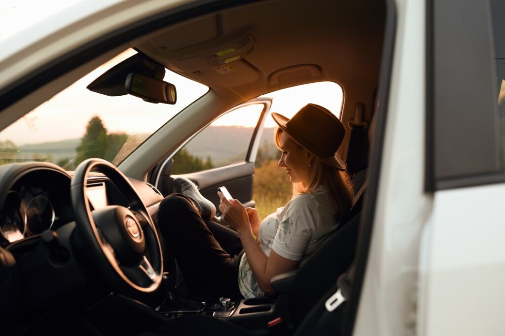 A woman checks her phone sitting the passenger seat of a car with the door open and the sunset light coming through.