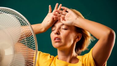 Woman standing directly in front of a fan with her hands up on her forehead trying to cool down.