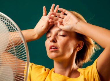 Woman standing directly in front of a fan with her hands up on her forehead trying to cool down.