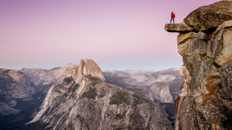 A man stands on a ledge over the epic view of Yosemite Valley which is a national park worth visiting at least once in your life.