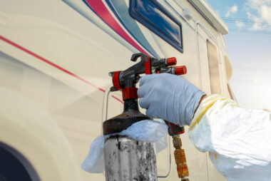 The hand of a person using a paint sprayer on the outside of an RV with red and blue decals