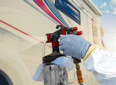 The hand of a person using a paint sprayer on the outside of an RV with red and blue decals