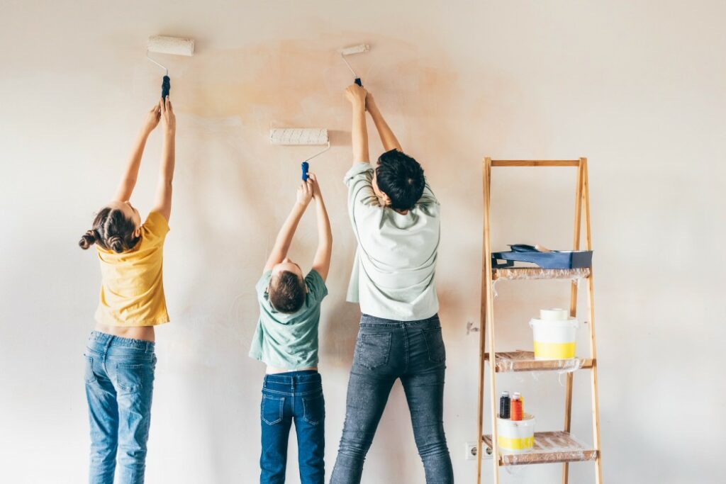 A family of three painting a wall with rollers and a ladder with supplies next to them.