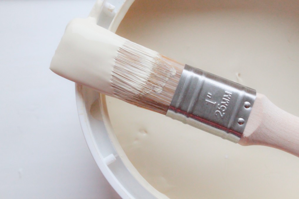 Close up of a small detail paint brush balanced on top of an open paint can with cream colored paint inside.